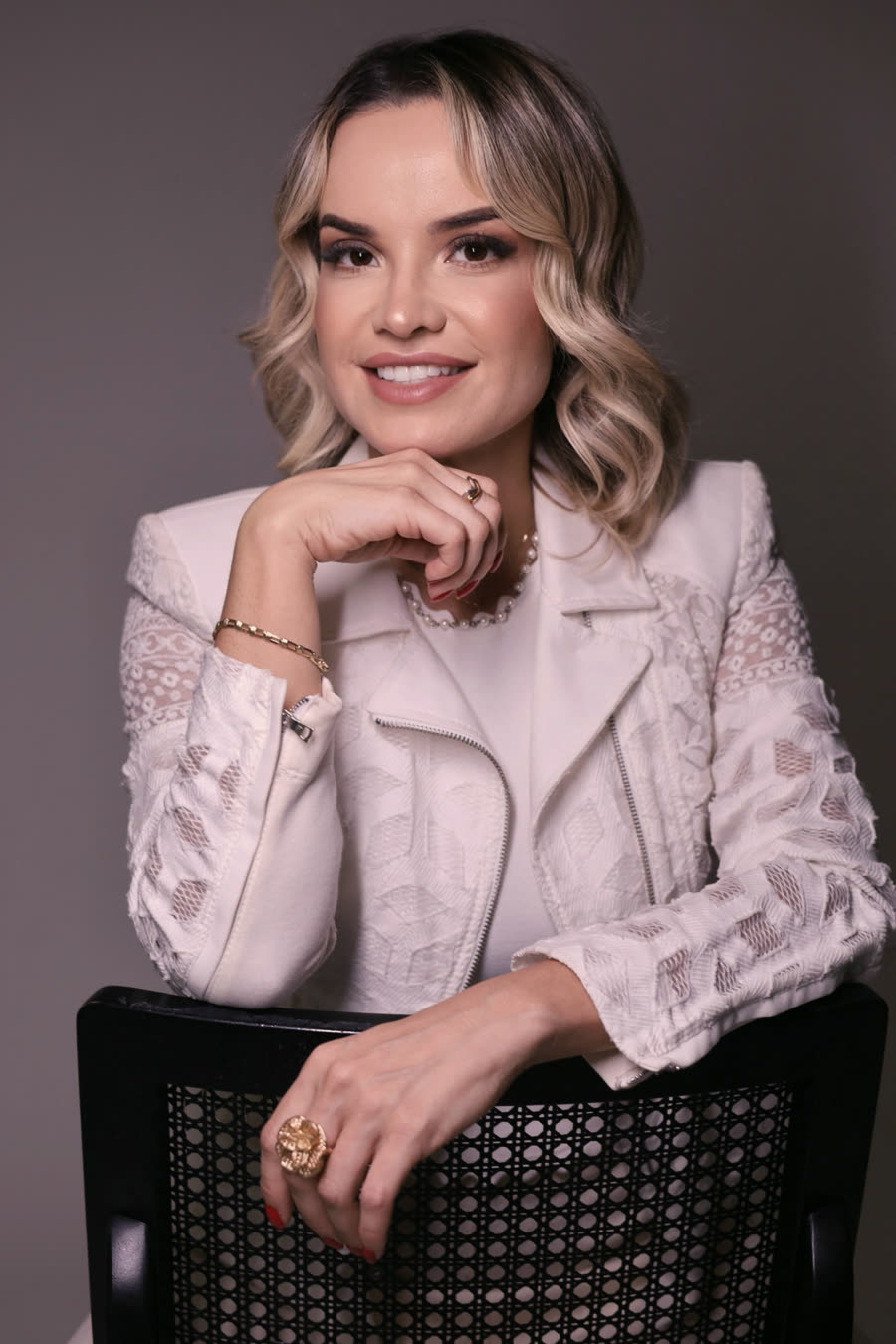 Pastora Andréa Castro sorrindo, de blazer branco rendado, apoiada no encosto de uma cadeira